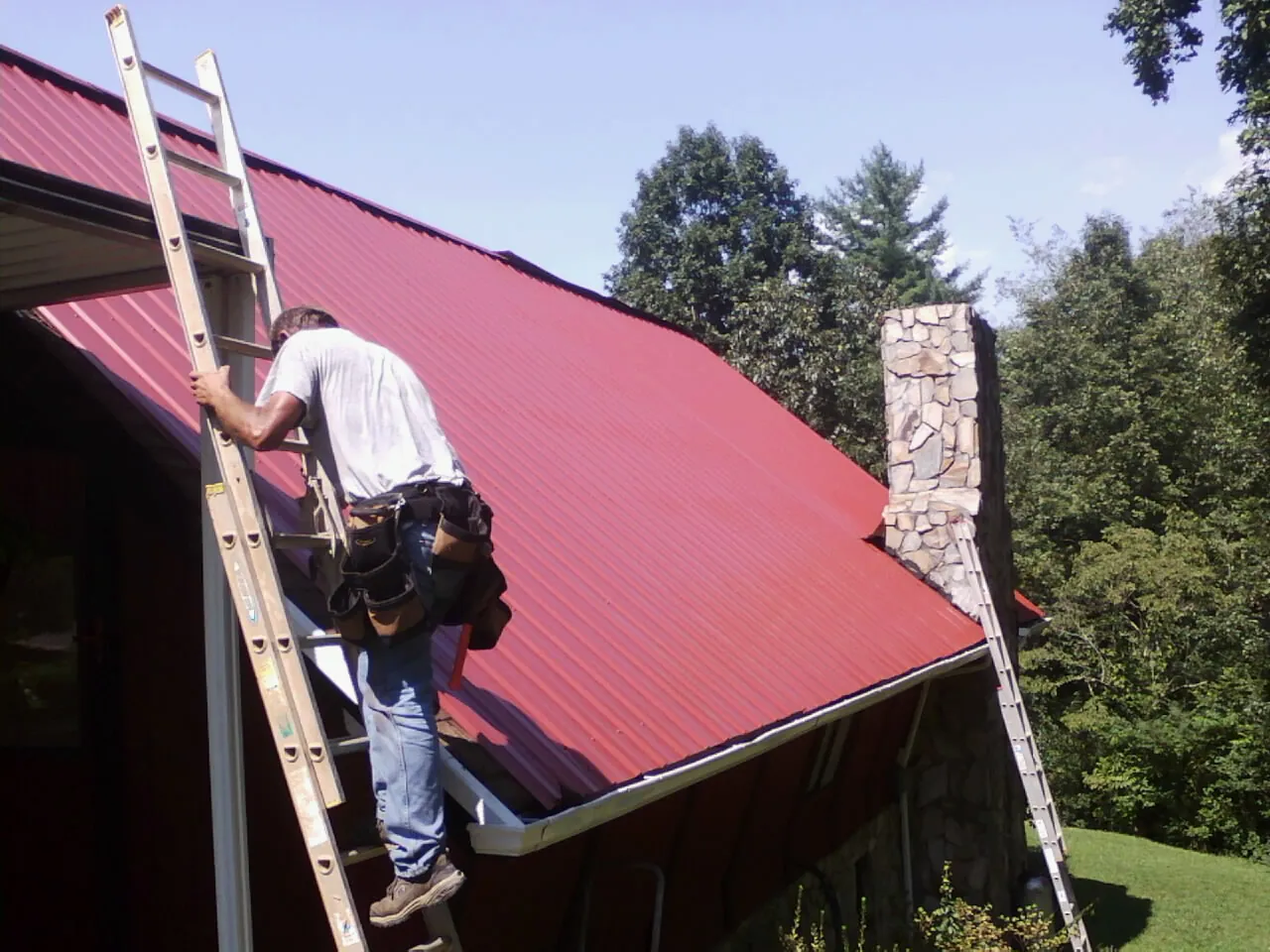 Roofer on a ladder beside a red metal roof and stone chimney — Soffit & Fascia Repair by Roofing Brothers in Fort Thomas